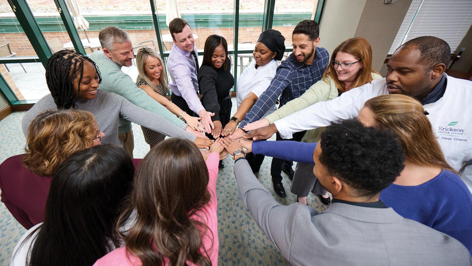 Diverse group of Erickson Senior Living professionals and staff standing in a circle with hands stacked together in a team-building gesture indoors.